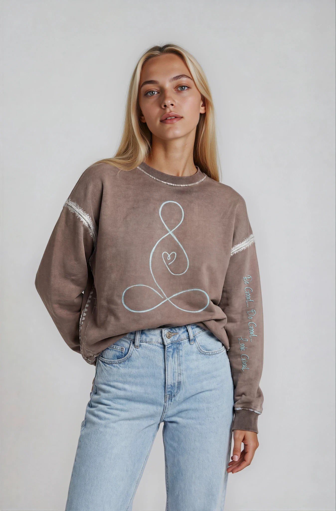Woman wearing a House-of-Malibu brown taupe sweatshirt with a painted buddha heart design and light blue jeans on a plain background